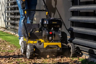 A person in blue jeans and white sneakers pushes a yellow and black Roll&Comb artificial grass sweeper next to a corrugated metal wall, clearing dry leaves and grass.