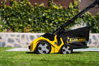 A yellow and black Garland Roll&Comb electric artificial turf sweeper with a black grass bag, shown on a green lawn with a blurred garden background.